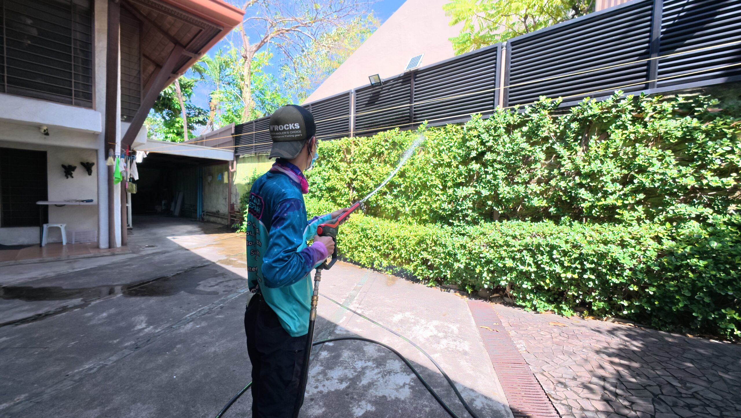 Professional exterior cleaner pre-watering a long green hedge along a driveway to protect the landscaping during house washing.